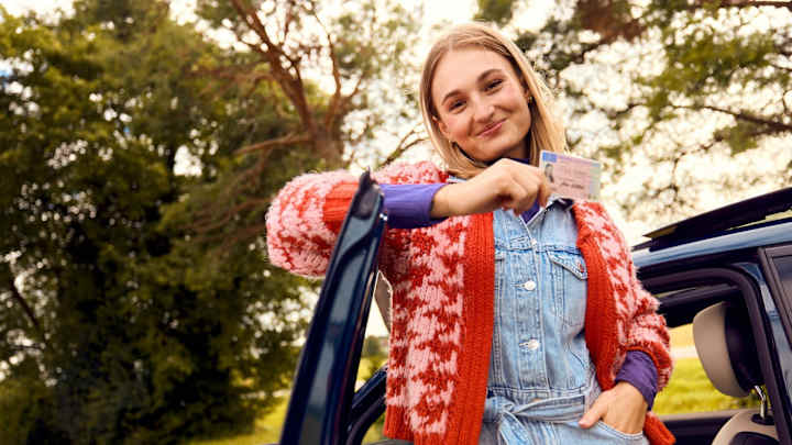 Junge Frau lehnt an der Autotüre mit ihrem Führerschein in der Hand