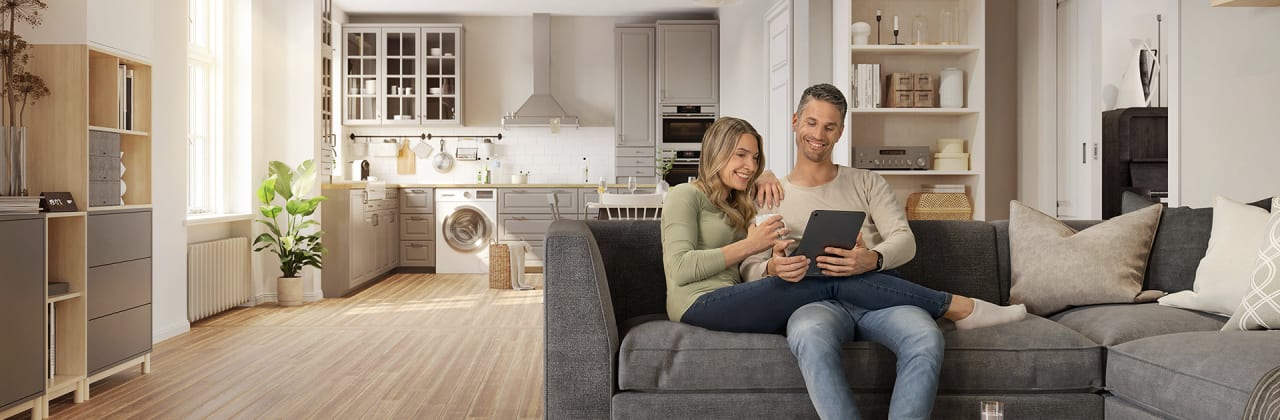 Junges Paar sitzt auf Sofa und schaut in ein Tablet, im Hintergrund Küche und Wohnzimmerschrank mit Hausrat