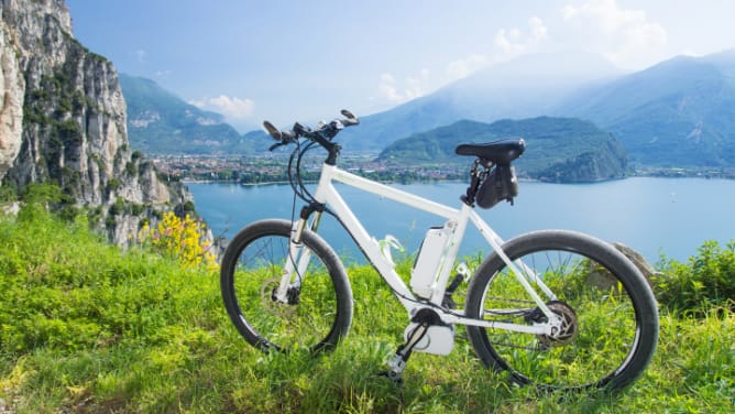 Weißes E-Bikes steht auf einem Hügel in der Wiese, im Hintergrund ein See und Berge