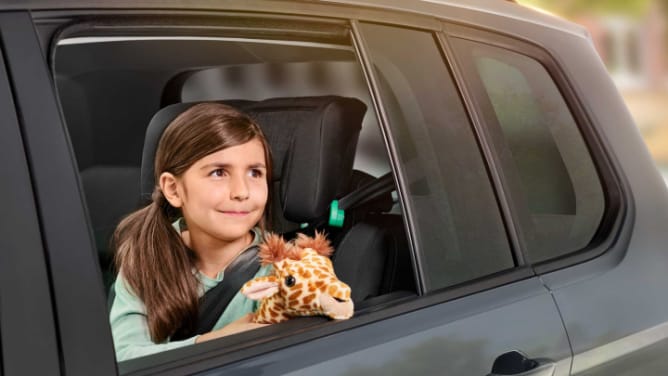 Maedchen sitzt mit Giraffe im Auto und lächelt aus dem Fenster - Autoversicherung ADAC