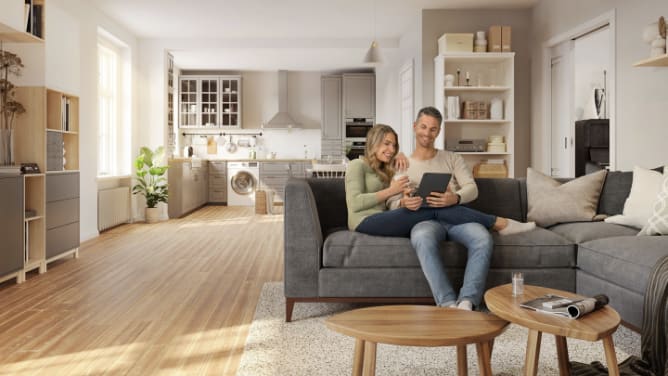 Junges Paar sitzt auf Sofa und schaut in ein Tablet, im Hintergrund Küche und Wohnzimmerschrank mit Hausrat