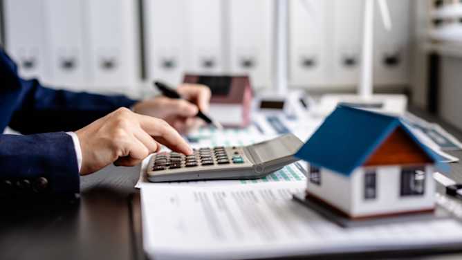 Tisch mit vielen Unterlagen, einem Modellhäuschen und einem Taschenrechner, auf dem jemand tippt