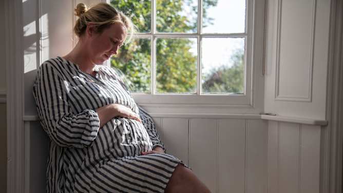 Reiserücktrittsversicherung Schwangerschaft: Frau sitzt am Fenster und schaut auf ihren Babybauch