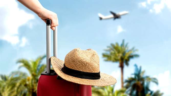Auslandskrankenversicherung: roter Koffer mit Sonnenhut im Vordergrund, im Hintergrund verschwommen Palmen und Flugzeit