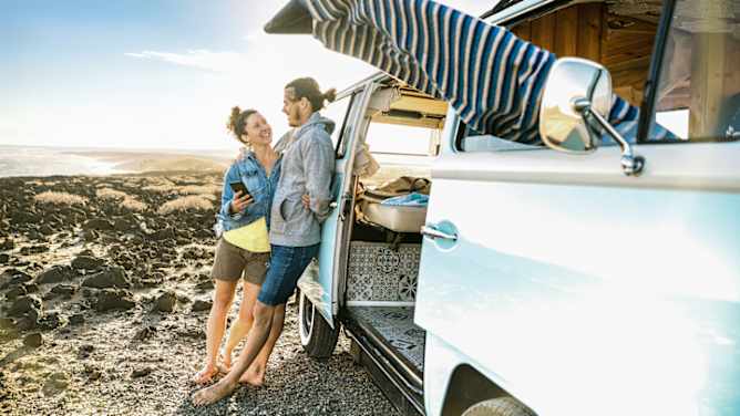 Auslandskrankenversicherung: Paar steht angelehnt am Bulli am Strand und lacht sich an
