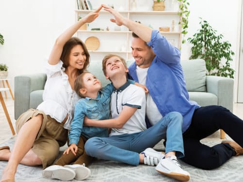 Familie sitzt vor der Couch auf dem Boden, Eltern heben Arme zu einem Dach über den Kindern