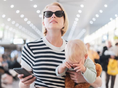 Mutter mit Kleinkind auf dem Arm steht am Flughafen und schaut auf Anzeigentafel