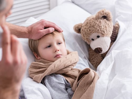 Kleiner Junge liegt krank im Bett, Vater fasst ihm an die Stirn während er telefoniert