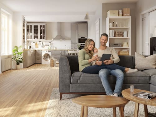 Junges Paar sitzt auf Sofa und schaut in ein Tablet, im Hintergrund Küche und Wohnzimmerschrank mit Hausrat