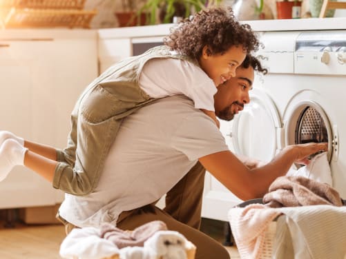 Mann mit Kind auf dem Rücken sitzt vor einer Waschmaschine