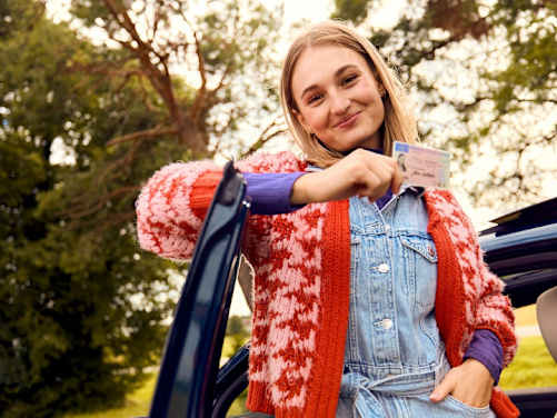 Junge Frau lehnt an der Autotüre mit ihrem Führerschein in der Hand