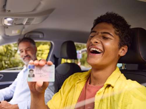 Junger Mann sitzt neben seinem Fahrlehrer im Auto und hält seinen Führerschein in der Hand
