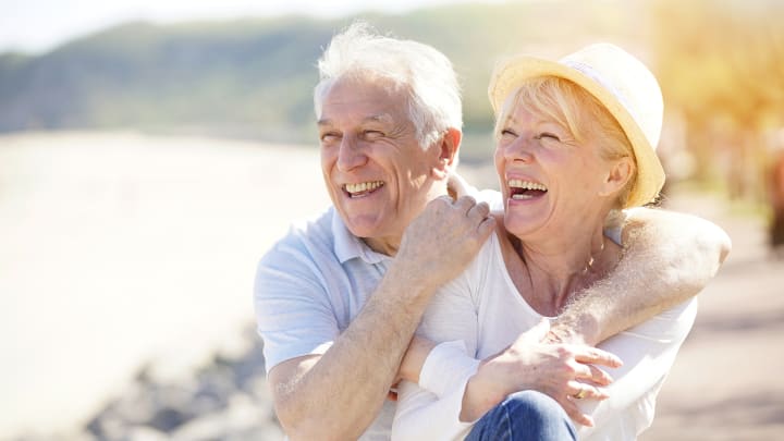 Älteres Paar schauen lachend und sich umarmend auf eine Bucht im Hintergrund: Auslandskrankenversicherung für Senioren