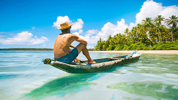 Mann sitzt in Longboat auf dem Meer und schaut auf einen Strand mit vielen Palmen