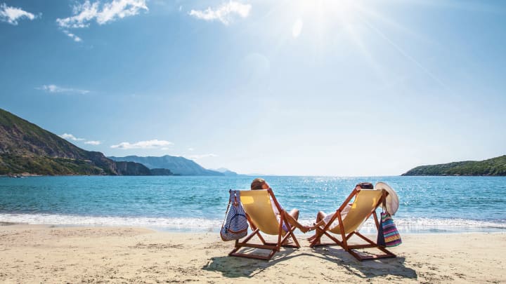 Reiseversicherungen: Paar sitzt am Strand in zwei Liegestühlen und hält dabei Händchen, vor ihnen das offene Meer