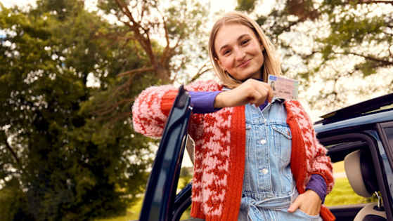 Junge Frau lehnt an der Autotüre mit ihrem Führerschein in der Hand