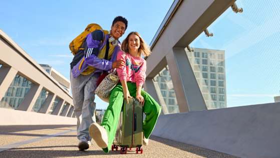 Zwei junge Leute sind mit Koffer und Taschen auf einer Brücke und lachen