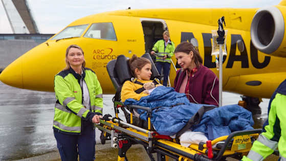 Kind liegt auf einer Krankenliege in Begleitung der Mutter mit dem ADAC Ambulanz-Jet im Hintergrund
