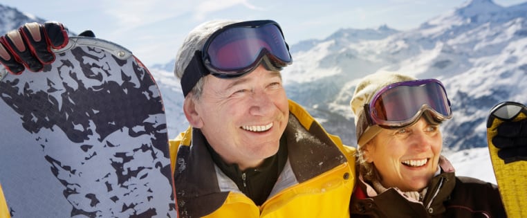 Senioren Paar steht mit Snowboard und Ski lachen auf der Skipiste - Unfallversicherung für Senioren