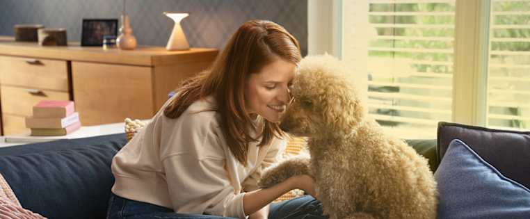 Frau sitzt mit Pudel auf einem Sofa und beugt ihren Kopf lächelnd zum Hund