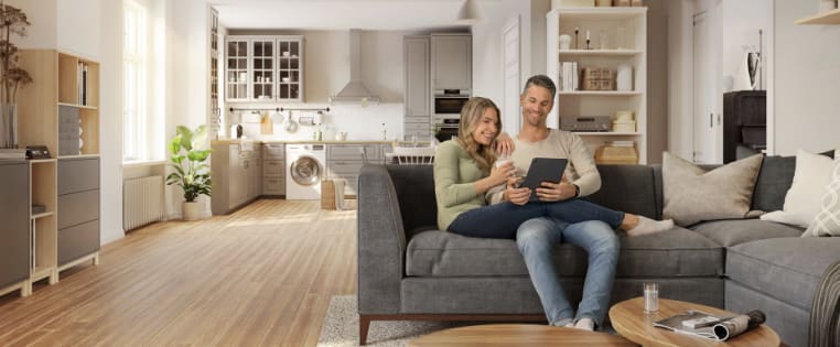 Junges Paar sitzt auf Sofa und schaut in ein Tablet, im Hintergrund Küche und Wohnzimmerschrank mit Hausrat