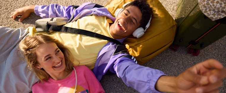 Zwei junge Leute lachen und liegen mit Tasche und Koffer auf dem Boden