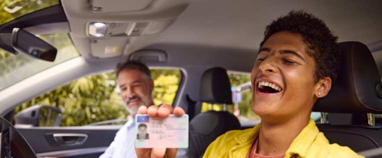 Junger Mann sitzt neben seinem Fahrlehrer im Auto und hält seinen Führerschein in der Hand