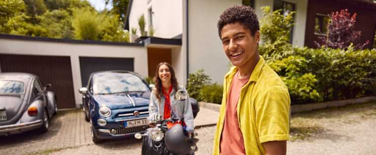 Zwei junge Leute mit Roller und Auto vor einer Hauseinfahrt