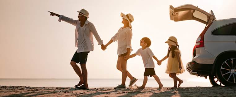 Familie läuft hintereinander am Strand entlang. Auto mit offenem Kofferraum - Kfz Versicherung Urlaub