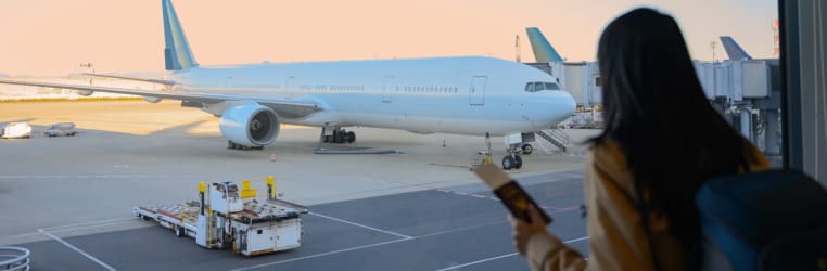 Frau steht mit Rucksack und Reisepass am Fenster des Flughafens und schaut auf ein Flugzeug