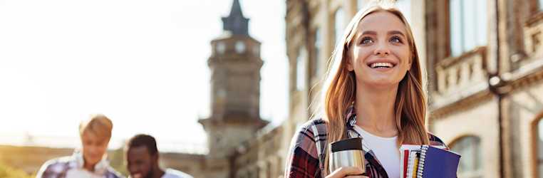 Junge Frau mit Kaffeebecher und Blöcken in der Hand steht vor einer alten Uni und schaut Richtung Himmel, im Hintergrund zwei junge Männer mit Tablet
