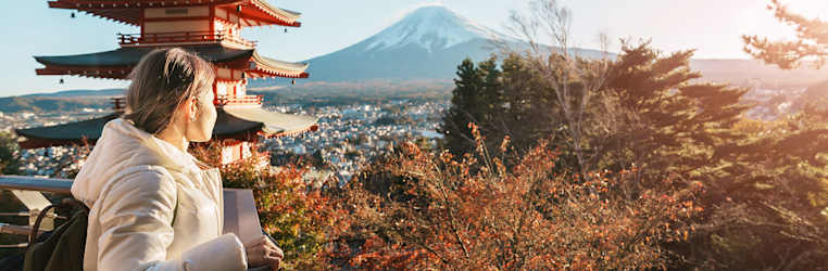 Junge Frau steht in Japan auf einer Aussichtsplatform und schaut weg von der Kamera auf einen schneebedeckten Berg, neben ihr ein Tempel